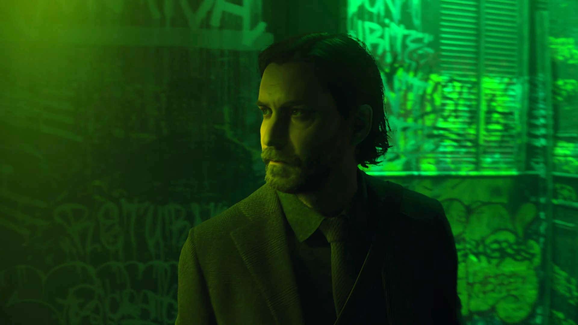 A screenshot of Alan Wake II shows Alan Wake standing in a green-lit alley.